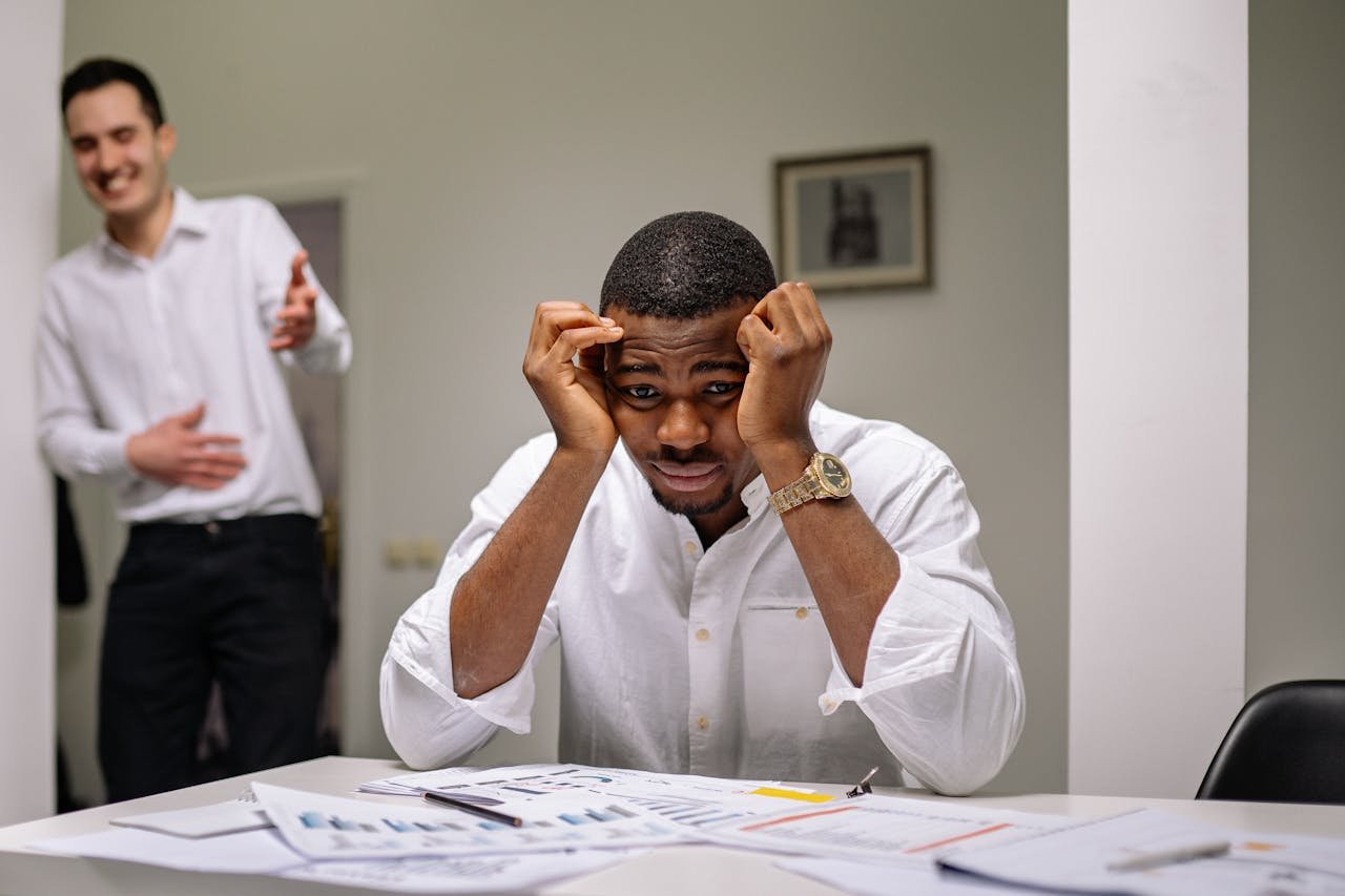 Tensed businessman at desk while coworker is laughing. Office stress concept.
